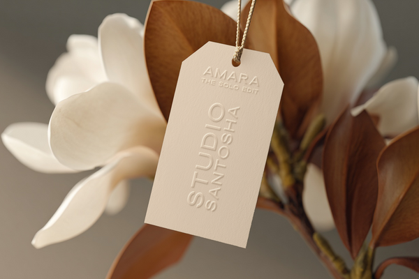 Beige vertical hang tag mockup featuring embossed ‘Studio Santosha’ and ‘Amara – The Solo Edit’ branding, suspended by a gold cord against magnolia flowers and warm brown leaves in soft natural light.