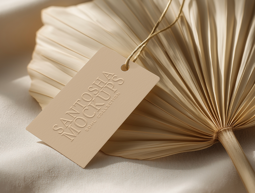 Hangtag mockup from the SOHL Collection by Studio Santosha, beige embossed tag styled on a dried palm leaf with soft fabric and natural light.